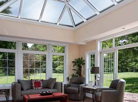 Interior of a sunlit sunroom with floor-to-ceiling windows and a peaked glass ceiling with the door open to a grassy yard with trees.